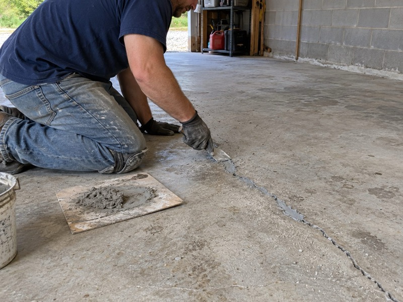 Person kneeling to trowel repair mortar into a long crack in a bare garage slab